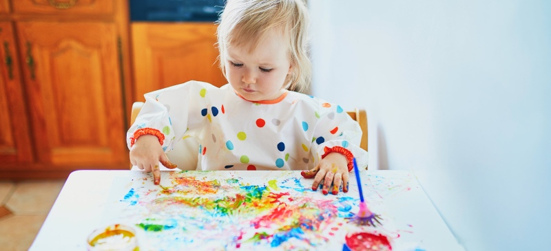Los niños, el juego y el arte. Niña jugando con pintura de dedos en una mesa, con la cara manchada.