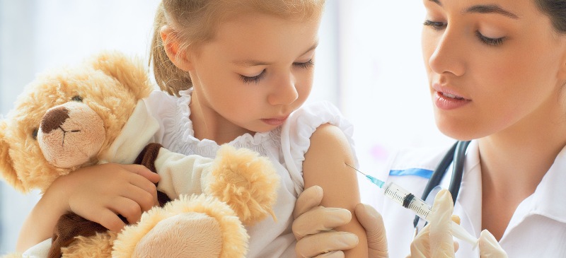 Vacunación de una niña con su osito de peluche y una enfermera que administra vacunas.