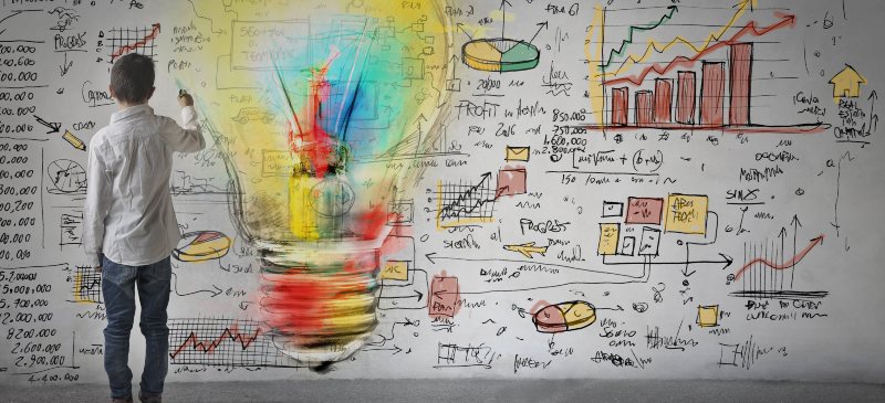 Niño de espaldas escribiendo sobre una pizarra llena de contenidos científicos entre los que destaca una bombilla dibujada en varios colores