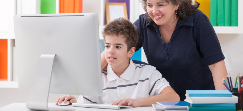 Alumno frente a un ordenador con una profesora por detrás observando lo que hace.