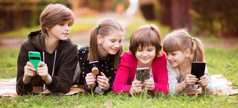 Las niñas en las TIC. Un grupo de niñas y niños mirando a sus teléfonos móviles sobre la hierba.