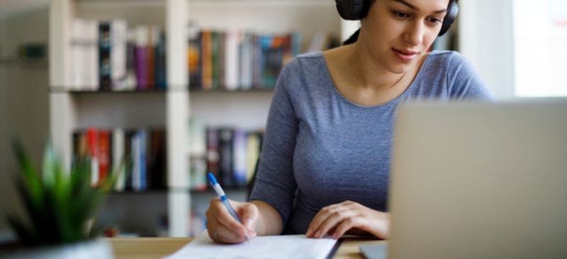 Chica estudiando un examen con cascos en su casa, preparando las oposiciones de Correos